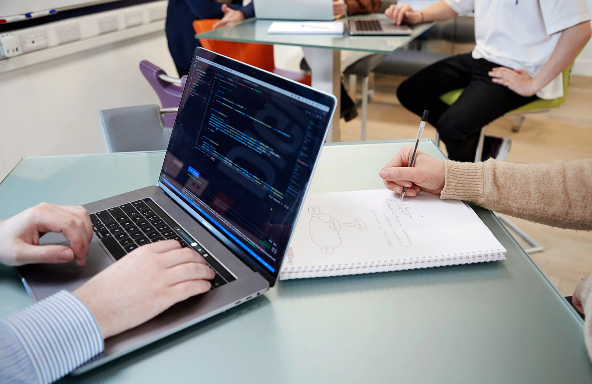 Two people working at a glass desk, one typing on a laptop displaying code and the other taking notes on a notepad, with others collaborating in the background.