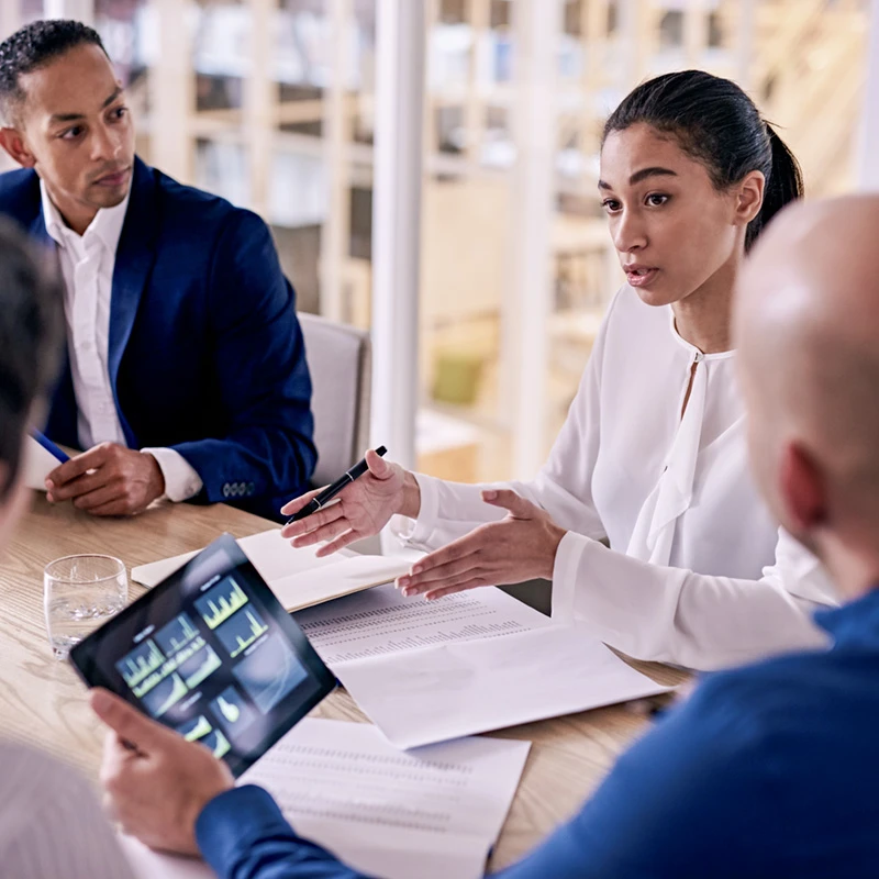 Woman presenting to colleagues in a meeting room, with papers and a tablet showing charts