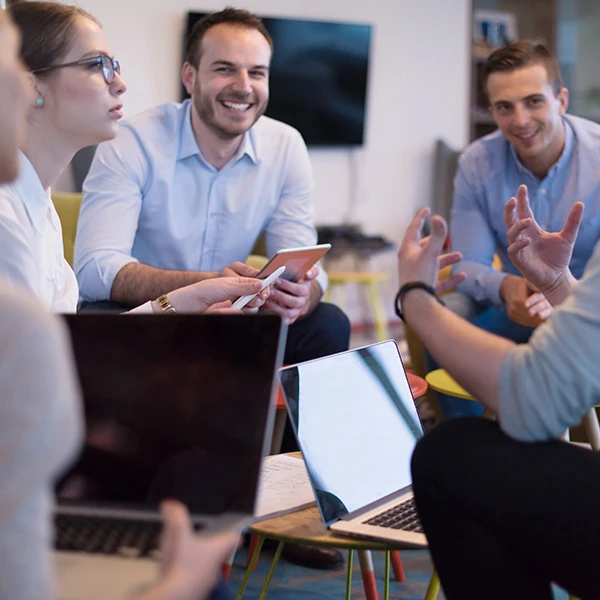 Group of professionals engaged in an informal meeting, sitting together with laptops and tablets, smiling and exchanging ideas, symbolising collaboration, teamwork, and open communication in a modern office setting.