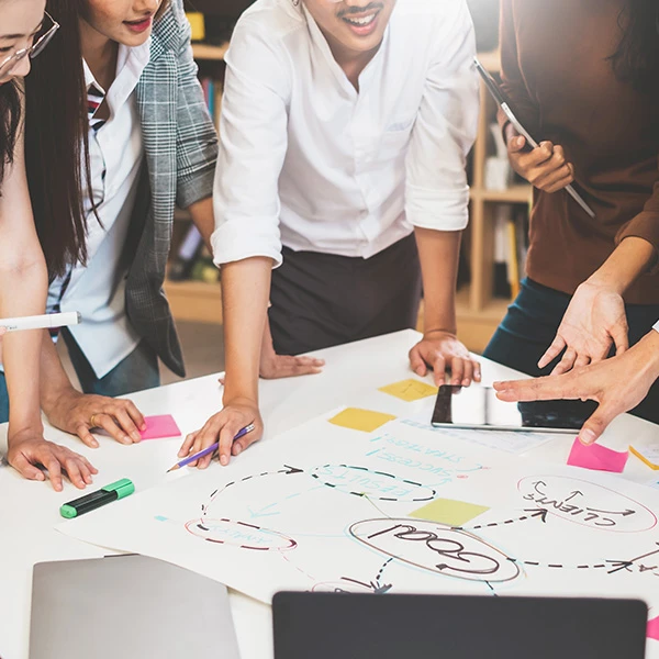 Team collaborating around a table, sketching a plan on a large sheet with arrows, markers and sticky notes.