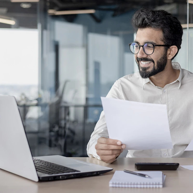 Smiling professional reviewing printed reports while working on a laptop in a modern glass office, suggesting successful project or financial results