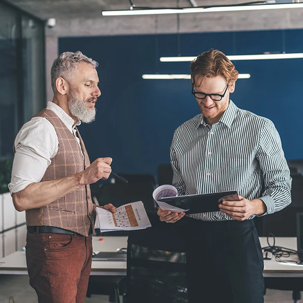 Two colleagues standing in a modern office discussing a document together, smiling as they review printed reports and project notes under bright lighting.