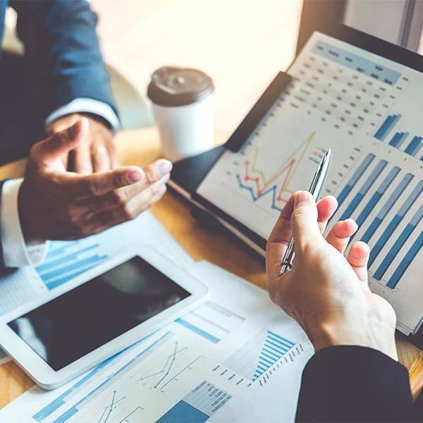 Close-up of a business meeting with hands gesturing over a laptop showing charts, papers with graphs, a tablet and a coffee cup on the table