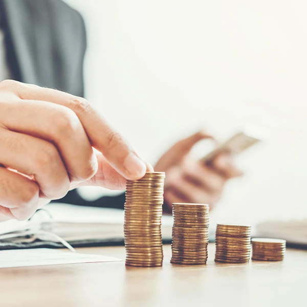 Close-up of a business person stacking coins into ascending piles on a desk, representing financial growth and savings, with a blurred figure holding a smartphone in the background.