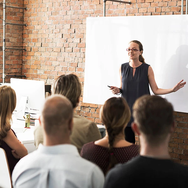 A woman leading a presentation to a small group in a modern office, standing in front of a whiteboard with a relaxed and confident posture, symbolising leadership, communication, and professional development.