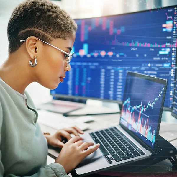 Focused professional analysing data charts and market trends on a laptop and dual monitors, with colourful graphs and performance metrics displayed across multiple screens.