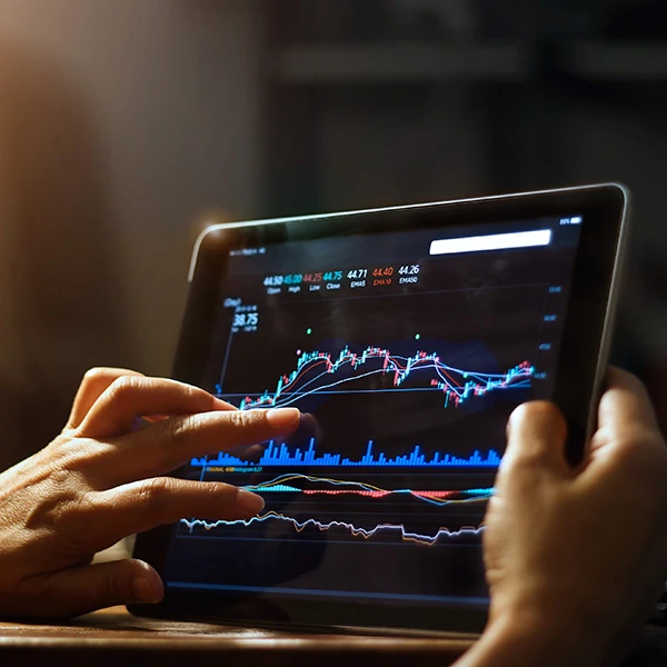 Hands using a tablet displaying a live stock market chart with indicators and volume bars.