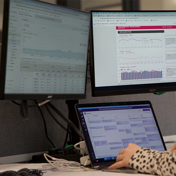 Desk setup with two monitors showing analytics dashboards and a laptop calendar, person typing