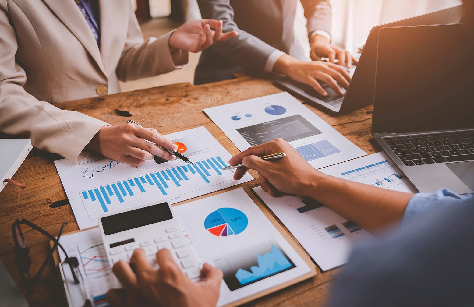 Business meeting scene showing a group of professionals reviewing financial charts, bar graphs, and reports on a wooden desk with laptops and calculators, representing financial analysis and strategic planning.