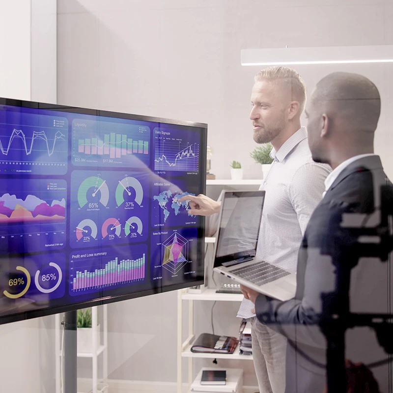 Two business professionals analysing performance metrics on a large digital display filled with graphs and data visualisations, while discussing insights with a laptop in hand