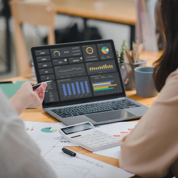 Two colleagues reviewing a laptop dashboard of KPIs, with a calculator and printed reports on the desk.