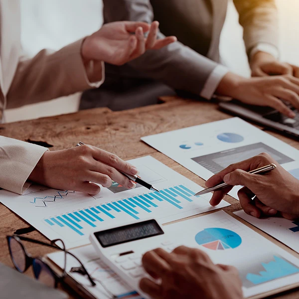 Group of professionals reviewing printed charts and digital data reports during a meeting, analysing performance metrics and discussing financial or marketing insights.