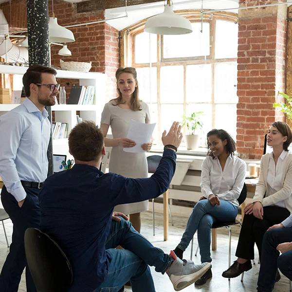 Team in a brick walled office during a leadership workshop, facilitator standing as participants discuss, one person raising a hand
