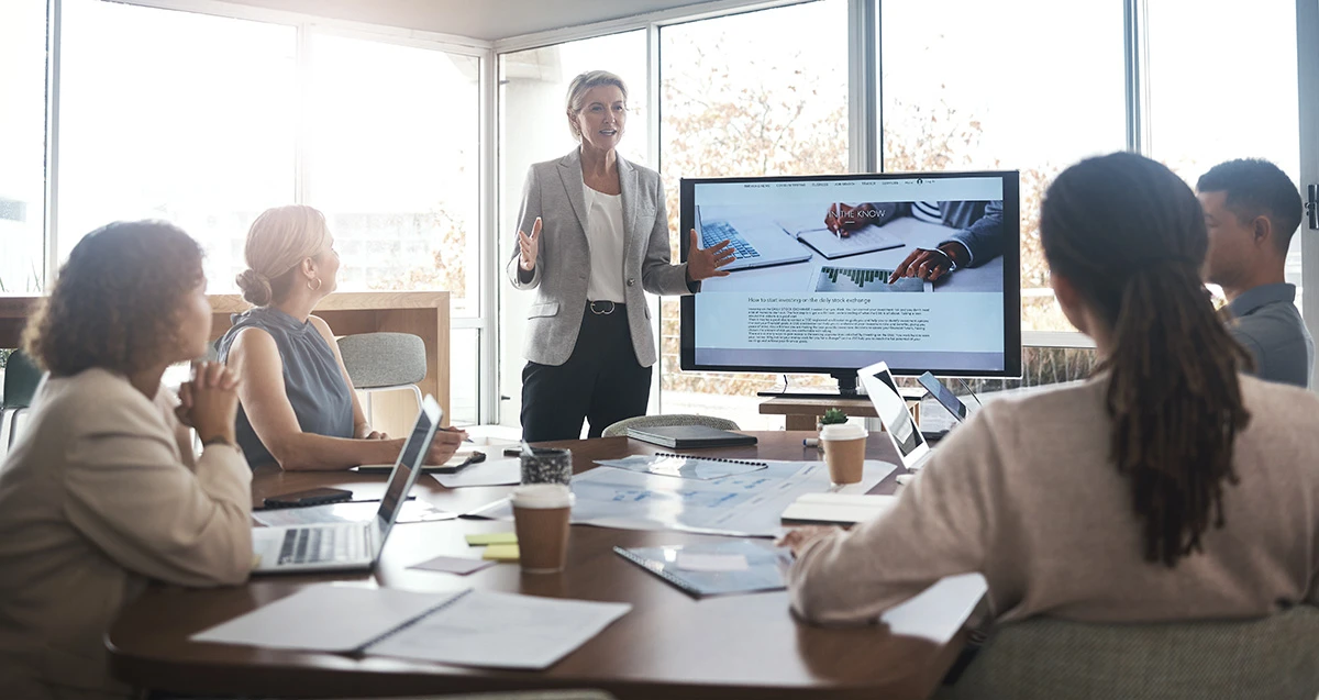 Executive presenting to a team in a bright boardroom, screen displaying charts, laptops on the table