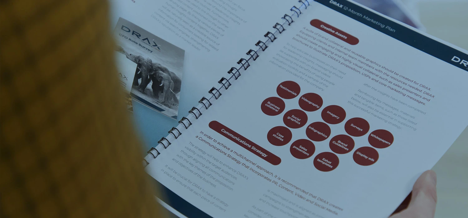 Person reading a printed marketing strategy document titled “DRAX 12-Month Marketing Plan,” featuring diagrams, red circular graphics, and structured text. The open booklet highlights sections on communications strategy and creative assets, symbolising professional marketing planning and strategy development.
