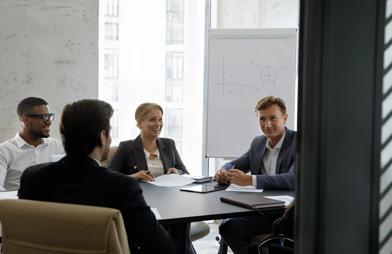 Business meeting with four professionals seated around a conference table, discussing strategy with charts and notes, and a whiteboard in the background displaying diagrams.