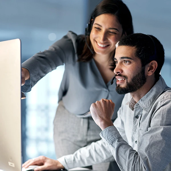 Two colleagues wearing headsets smiling as they collaborate at a computer, with one pointing at the screen and the other attentively following along, representing teamwork, system training, and digital onboarding.