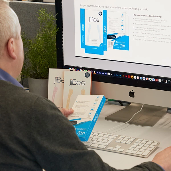 Designer reviewing JiBee product packaging designs on a computer screen while holding physical packaging boxes, checking updated artwork and layout details in an office setting.