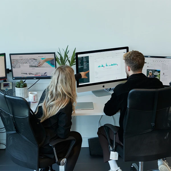 Two colleagues collaborating at a workstation with multiple computer monitors, analysing digital dashboards and data visualisations on screen. One person is pointing at a chart, while the other observes attentively, representing data reporting, digital marketing analysis, and performance review.