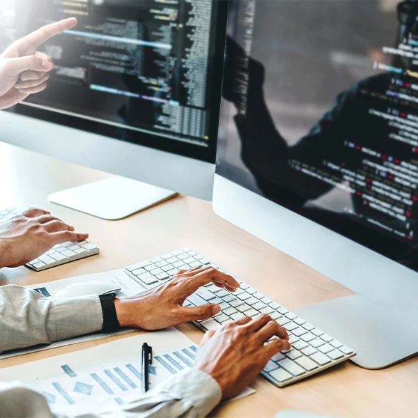 Developers working at desktop computers, hands on keyboards with code on screen and a colleague pointing at the monitor.