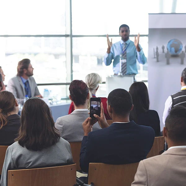 Audience attending a business seminar where a speaker presents on stage, with one participant recording the session on their phone.