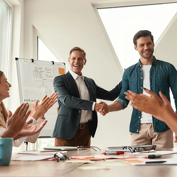 Team celebrating a staff achievement as two colleagues shake hands in a bright meeting room, surrounded by clapping colleagues and presentation charts in the background